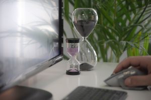 Hourglass on a table while person is working on a computer to demonstrate a guide to time management